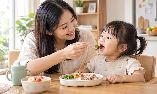 Seorang ibu sedang mendampingi balita yang belajar makan sendiri dengan tenang dan menyenangkan di meja makan