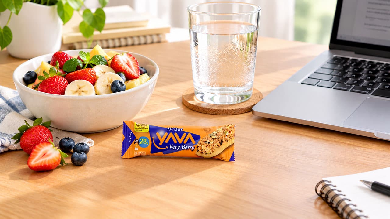 YA’Bar dan Buah untuk Camilan di Sela Aktivitas YA’Bar, buah segar, dan segelas air putih di atas meja kerja di samping laptop.